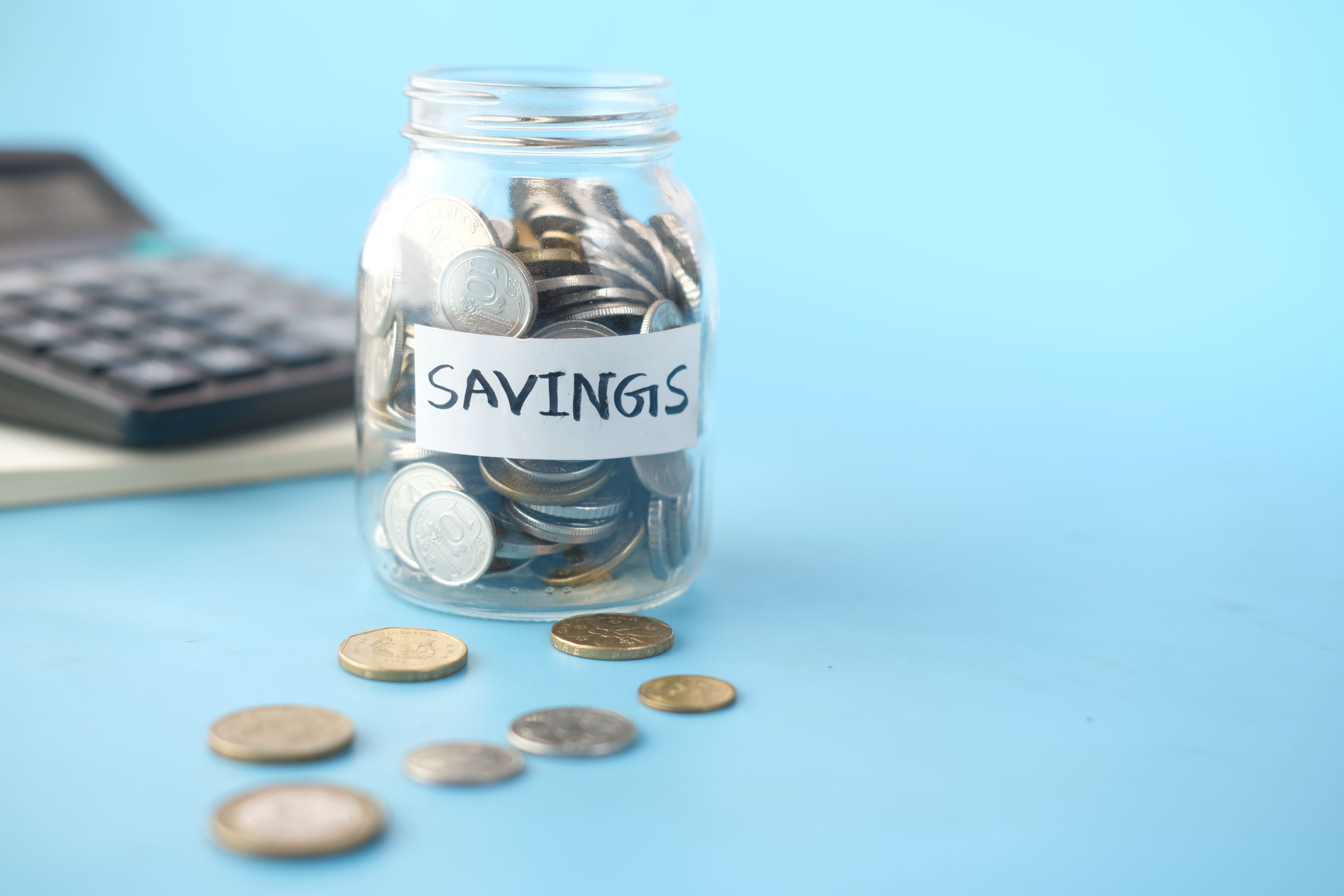 Glass savings jar filled with coins beside a calculator on a blue background.